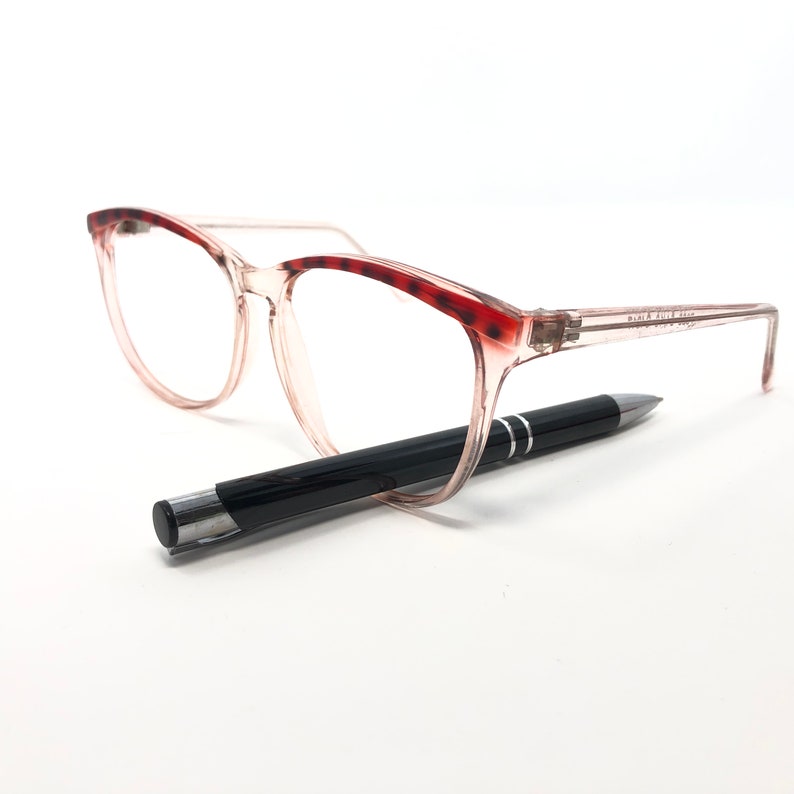 May include: Clear-framed eyeglasses with red tortoise shell accents on the upper rims. A black pen with silver accents rests beneath the glasses. The glasses have a rectangular shape.