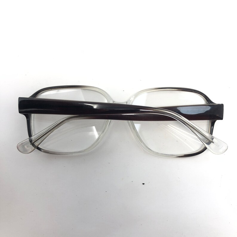 K&ouml;nnte beinhalten: Eine Brille mit transparentem Rahmen und dunkelbraunen B&uuml;geln. Die Brille hat eine rechteckige Form. Die Aufnahme zeigt die Brille von oben, wobei die Gl&auml;ser und das Design des Rahmens hervorgehoben werden.