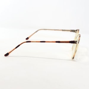 K&ouml;nnte beinhalten: Eine Brille mit einem transparenten, runden Rahmen und d&uuml;nnen, silberfarbenen Metallb&uuml;geln. Die B&uuml;gel sind hellbraun mit dunkelbraunen Akzenten. Die Gl&auml;ser sind klar, vor wei&szlig;em Hintergrund.
