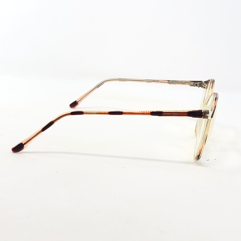 K&ouml;nnte beinhalten: Eine Brille mit einem transparenten, runden Rahmen und d&uuml;nnen, silberfarbenen Metallb&uuml;geln. Die B&uuml;gel sind hellbraun mit dunkelbraunen Akzenten. Die Gl&auml;ser sind klar, vor wei&szlig;em Hintergrund.