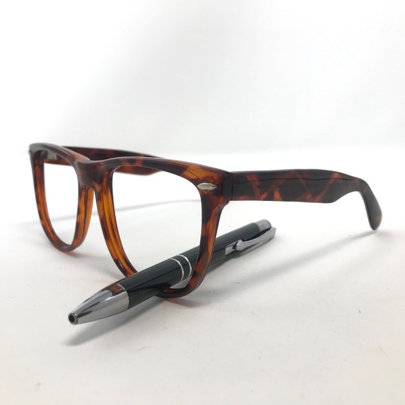 May include: A pair of tortoiseshell eyeglasses with a black pen resting beneath the frames. The glasses have a classic, rectangular shape and are made of a brown and amber-colored material. The pen is black with silver accents.