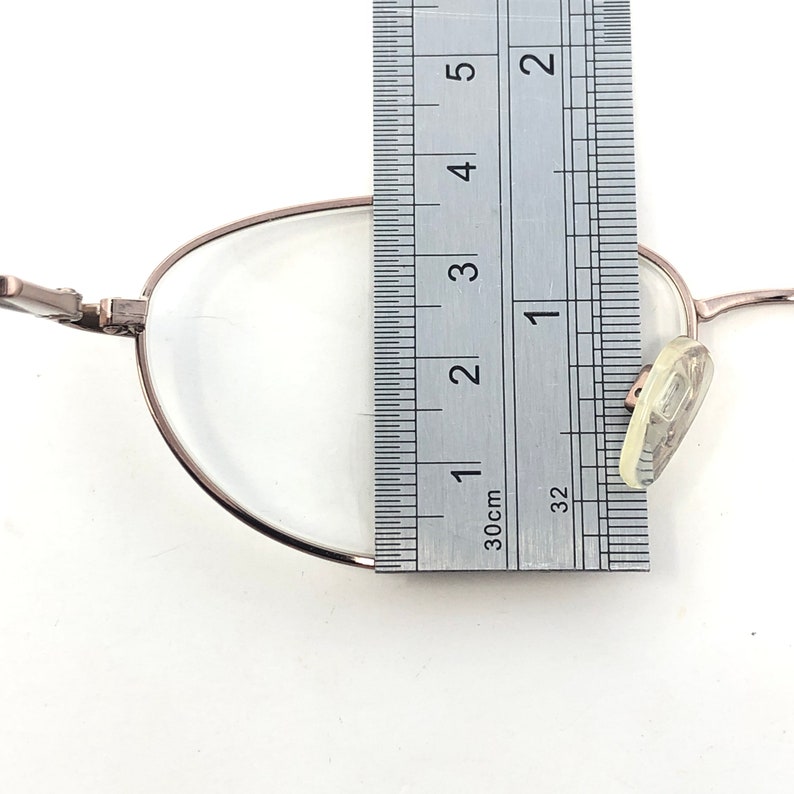 May include: Silver-framed eyeglasses with clear lenses, measured with a ruler. The ruler shows a measurement of approximately 1 inch. The glasses have nose pads and are set against a white background.