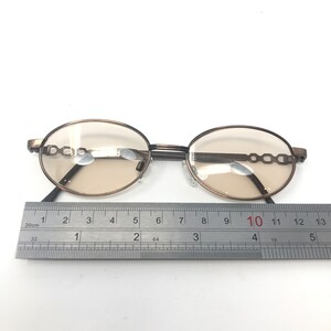 May include: Oval-shaped eyeglasses with a bronze-colored frame and tinted lenses. The frame features a decorative chain-link design on the temples. A ruler is placed below the glasses, measuring approximately 4 inches in length.