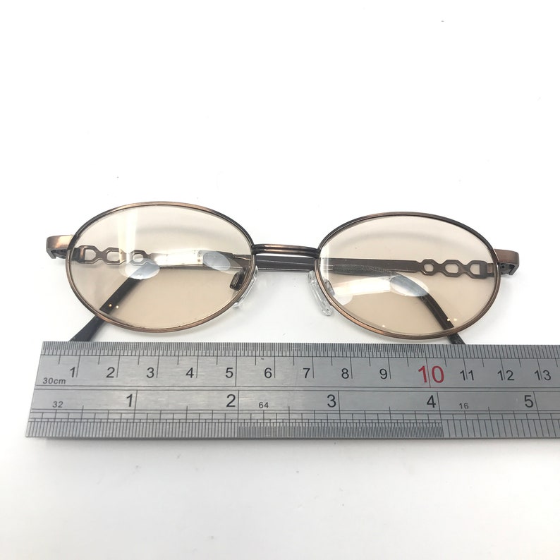 May include: Oval-shaped eyeglasses with a bronze-colored frame and tinted lenses. The frame features a decorative chain-link design on the temples. A ruler is placed below the glasses, measuring approximately 4 inches in length.