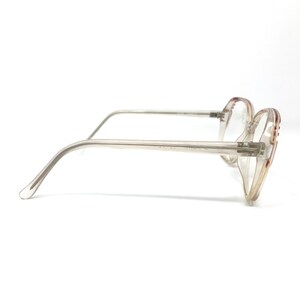 May include: Clear-framed eyeglasses with a light brown tint and red accents on the temples. The glasses have a classic, retro design with a rectangular shape. The arms are thin and transparent, and the lenses are clear.