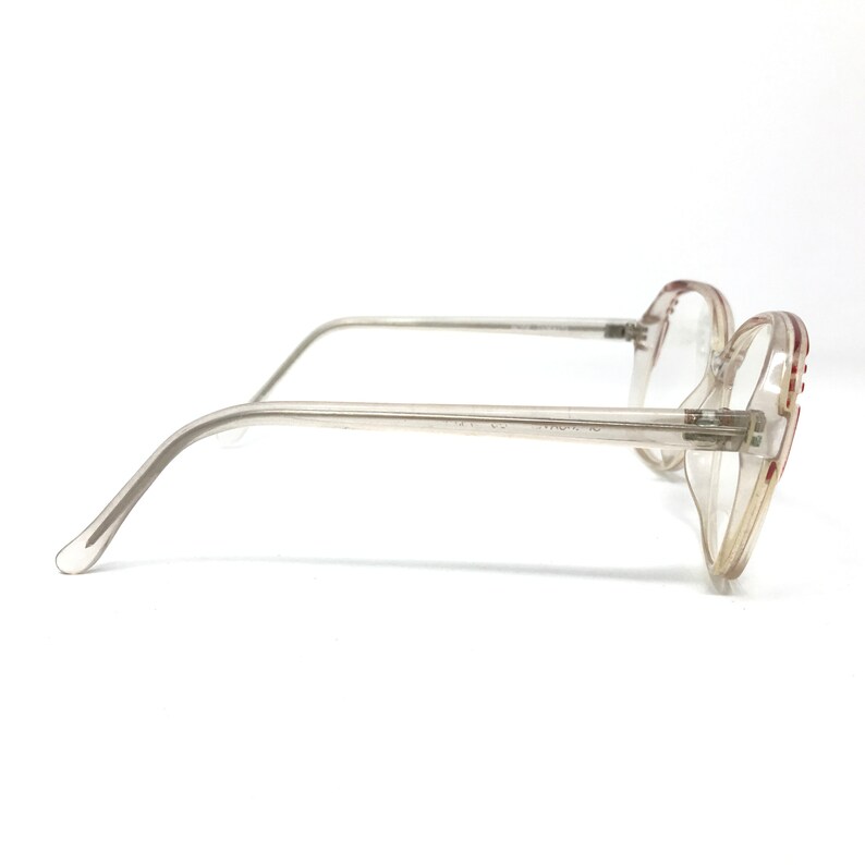 May include: Clear-framed eyeglasses with a light brown tint and red accents on the temples. The glasses have a classic, retro design with a rectangular shape. The arms are thin and transparent, and the lenses are clear.