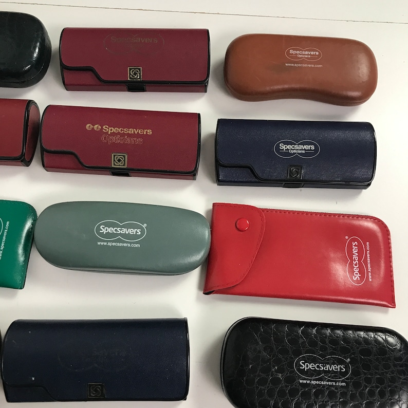May include: A collection of eyeglass cases in various colors, including red, blue, and black. The cases have the "Specsavers" logo printed on them. Some cases are rectangular, while others are oval-shaped.