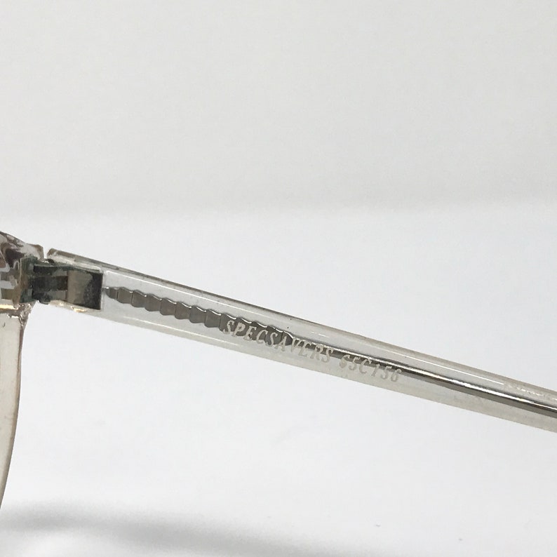 Puede incluir: Primer plano de una montura de gafas transparente con el texto "SPECSAVERS SSC756" impreso en la varilla. La montura es de material transparente y tiene forma rectangular.