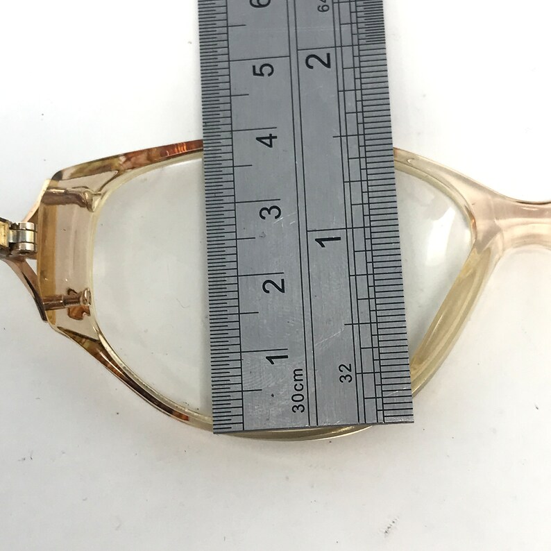 May include: A pair of eyeglasses with a light brown frame, measured with a ruler. The ruler shows the lens is approximately 1.5 inches wide. The glasses have a rectangular shape and clear lenses.