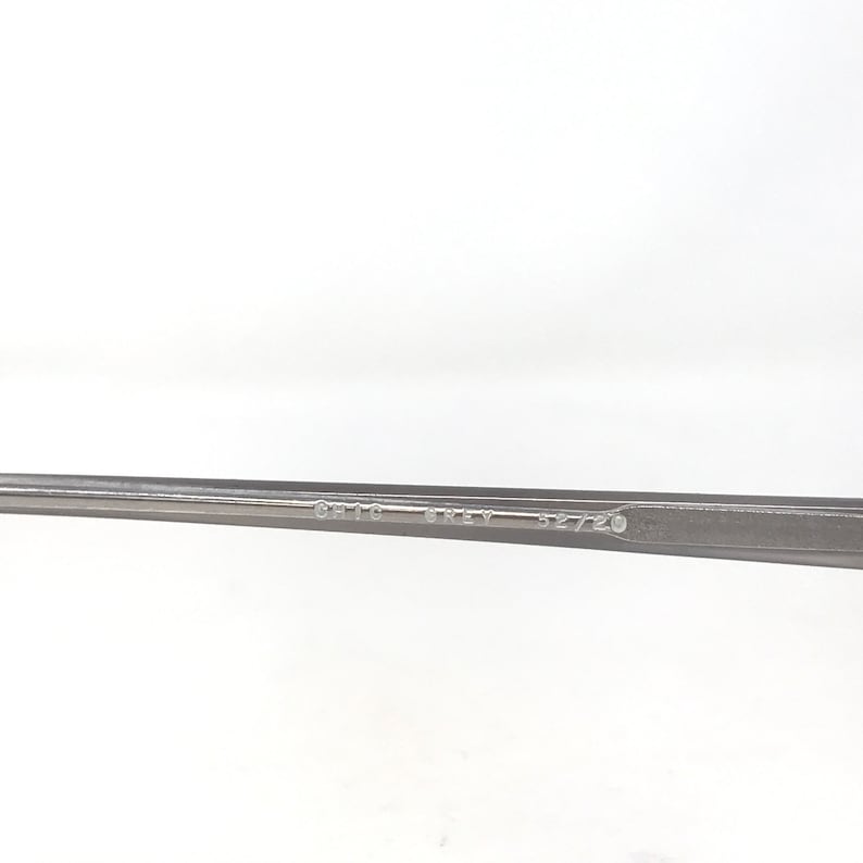 May include: A close-up of a gray metal eyeglass frame. The text "CHIC GREY 52/20" is printed on the frame. The frame is set against a white background.