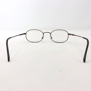 Puede incluir: Gafas de forma ovalada con montura de metal marr&oacute;n oscuro. Las lentes son transparentes y las patillas tienen terminales negros. Las gafas est&aacute;n sobre un fondo blanco.