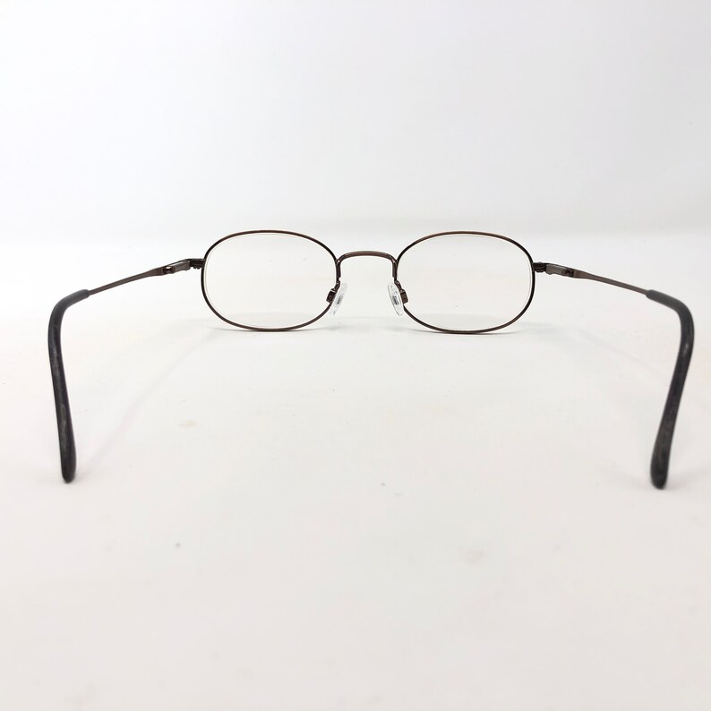 Puede incluir: Gafas de forma ovalada con montura de metal marr&oacute;n oscuro. Las lentes son transparentes y las patillas tienen terminales negros. Las gafas est&aacute;n sobre un fondo blanco.