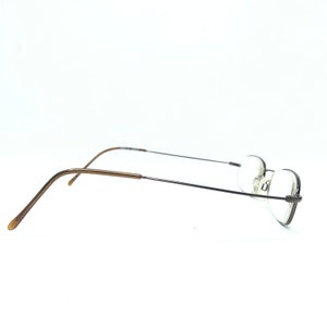 Puede incluir: Gafas sin montura con una fina montura de metal oscuro y lentes ovaladas. Las patillas son de color marr&oacute;n claro. Las gafas est&aacute;n sobre un fondo blanco.