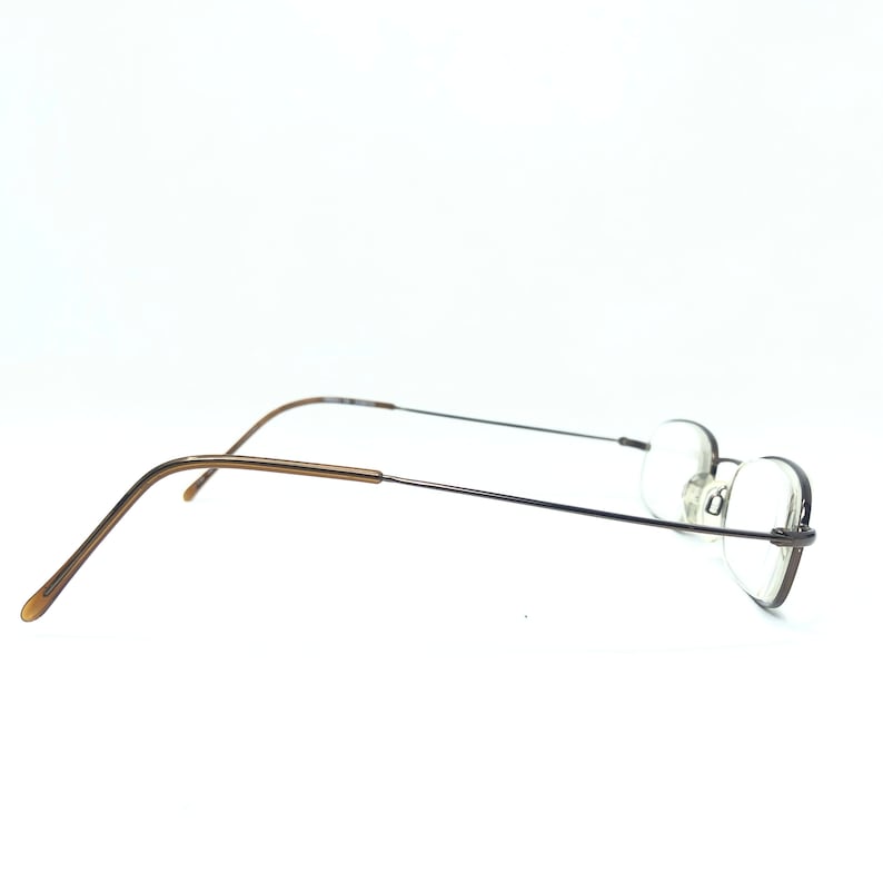 Puede incluir: Gafas sin montura con una fina montura de metal oscuro y lentes ovaladas. Las patillas son de color marr&oacute;n claro. Las gafas est&aacute;n sobre un fondo blanco.