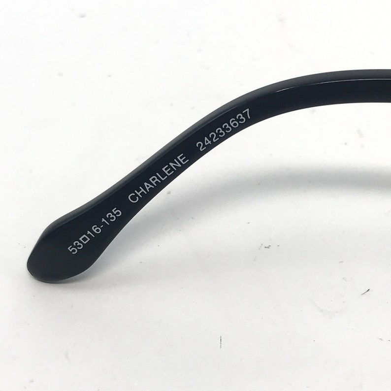 Puede incluir: Primer plano de la patilla de una montura de gafas negra. El texto "53▢16-135 CHARLENE 2423637" est&aacute; impreso en blanco en la patilla. La montura est&aacute; sobre un fondo blanco.