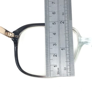 May include: Black-framed eyeglasses with a clear lens are measured with a ruler. The frame is approximately 5 cm wide. The glasses have a gold-coloured hinge and are set against a white background.