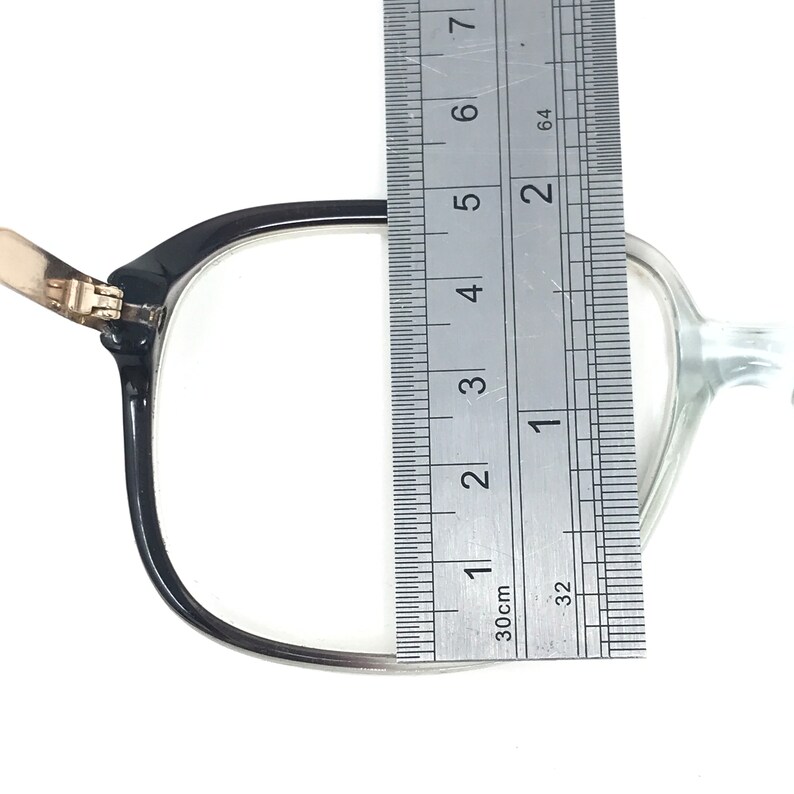 May include: Black-framed eyeglasses with a clear lens are measured with a ruler. The frame is approximately 5 cm wide. The glasses have a gold-coloured hinge and are set against a white background.