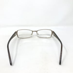 May include: Rectangular eyeglasses with a silver metal frame and dark brown temples. The glasses are set against a white background, showcasing their design and structure. The lenses are clear, and the overall aesthetic is modern and functional.