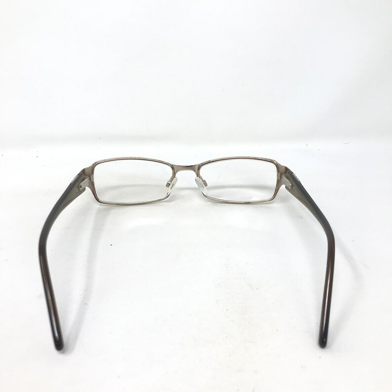 May include: Rectangular eyeglasses with a silver metal frame and dark brown temples. The glasses are set against a white background, showcasing their design and structure. The lenses are clear, and the overall aesthetic is modern and functional.