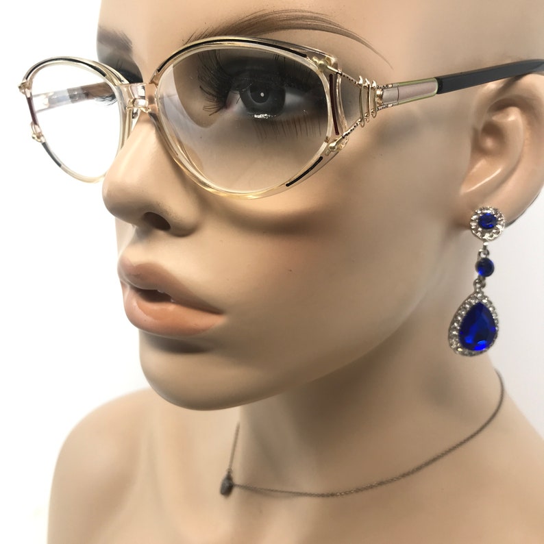 Puede incluir: Gafas con montura transparente, un acento dorado y patillas negras. El modelo tambi&eacute;n lleva un par de pendientes azules en forma de l&aacute;grima y un collar con colgante a juego. Las gafas tienen un estilo retro.