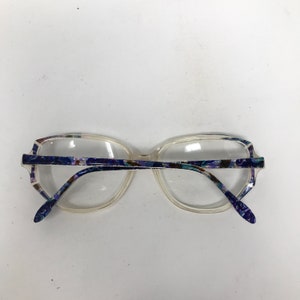Puede incluir: Gafas transparentes con montura rectangular y estampado floral en las patillas. La montura es de color claro y las patillas tienen un dise&ntilde;o floral azul, morado y verde. Las lentes son transparentes.