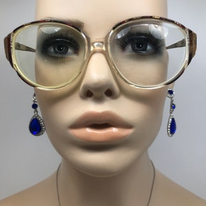 May include: Clear, oversized eyeglasses with a light brown frame and gold accents. The glasses have a rectangular shape with rounded edges. The image also shows a pair of blue teardrop earrings.