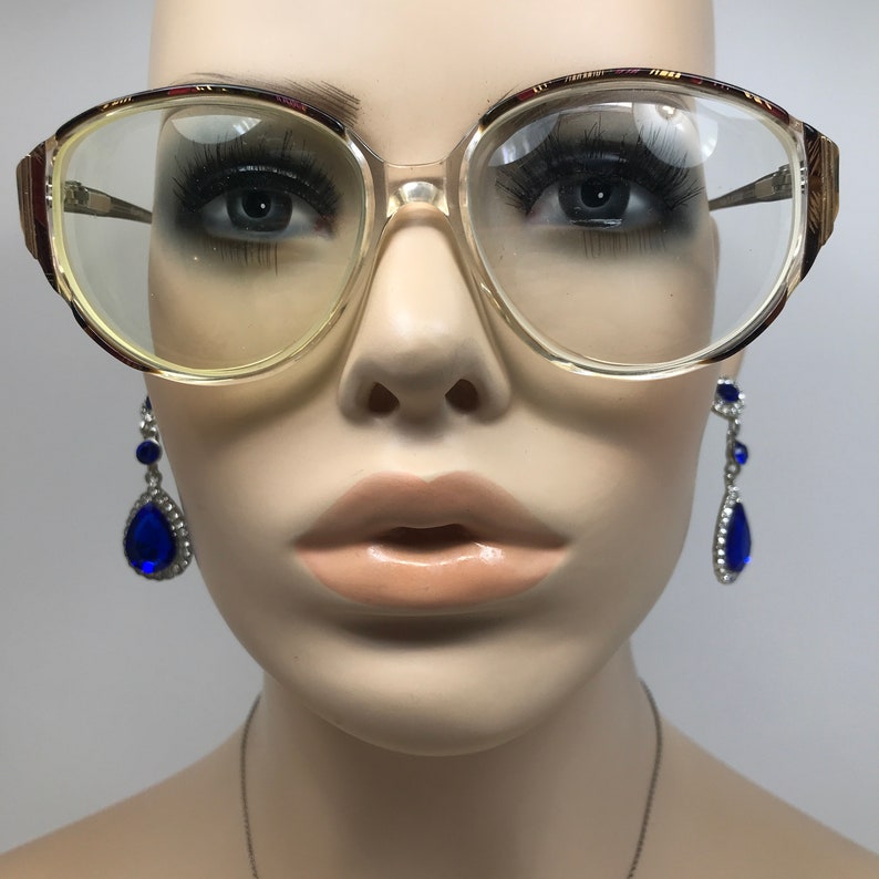 May include: Clear, oversized eyeglasses with a light brown frame and gold accents. The glasses have a rectangular shape with rounded edges. The image also shows a pair of blue teardrop earrings.
