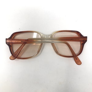 May include: Vintage eyeglasses with a gradient brown and clear frame. The glasses have a rectangular shape and are viewed from the back, showcasing the arms and the lenses. The lenses have a light tint.