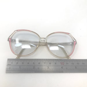 May include: Clear eyeglasses with a pink accent on the frame. The glasses have a large, round shape. A ruler is placed below the glasses, measuring up to 14 inches.