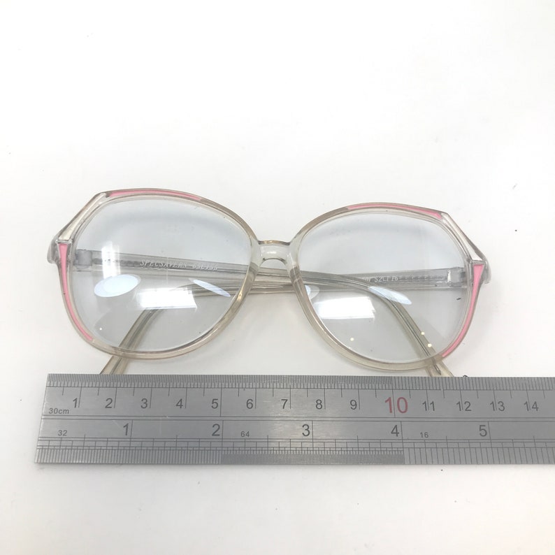 May include: Clear eyeglasses with a pink accent on the frame. The glasses have a large, round shape. A ruler is placed below the glasses, measuring up to 14 inches.