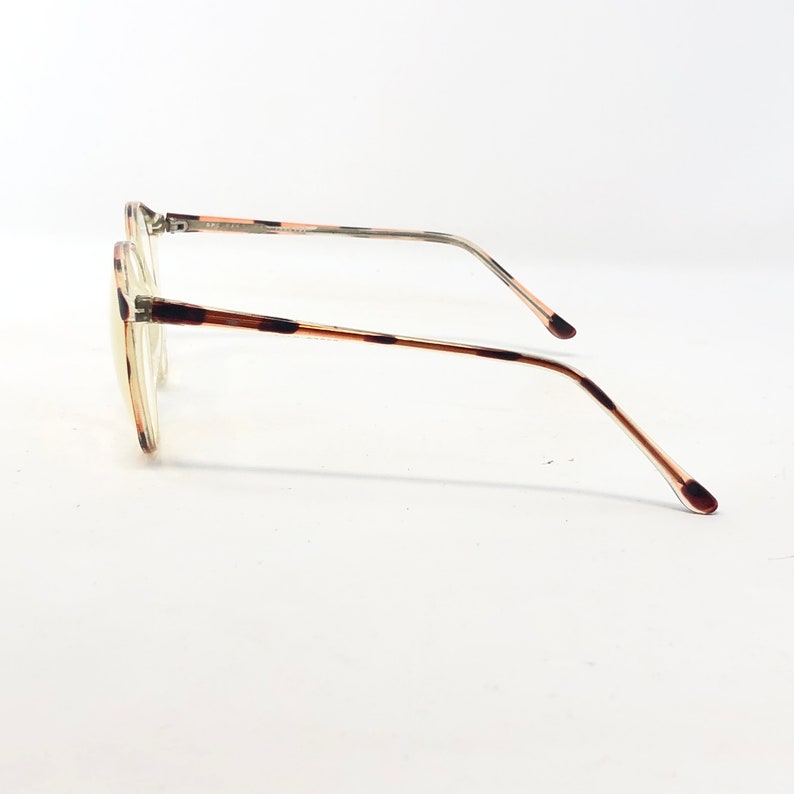 K&ouml;nnte beinhalten: Eine Brille mit hellbraunem Schildpattmuster auf dem Rahmen und den B&uuml;geln. Die Gl&auml;ser sind klar, und die B&uuml;gel haben ein d&uuml;nnes, gebogenes Design. Die Brille ist vor einem wei&szlig;en Hintergrund platziert.