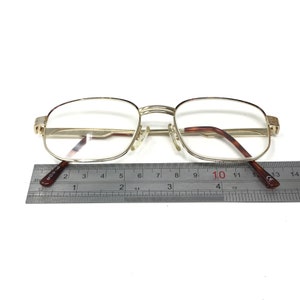 May include: Gold-tone metal eyeglasses with a rectangular frame and clear lenses. The eyeglasses are measured with a ruler, showing a width of approximately 10 centimeters.