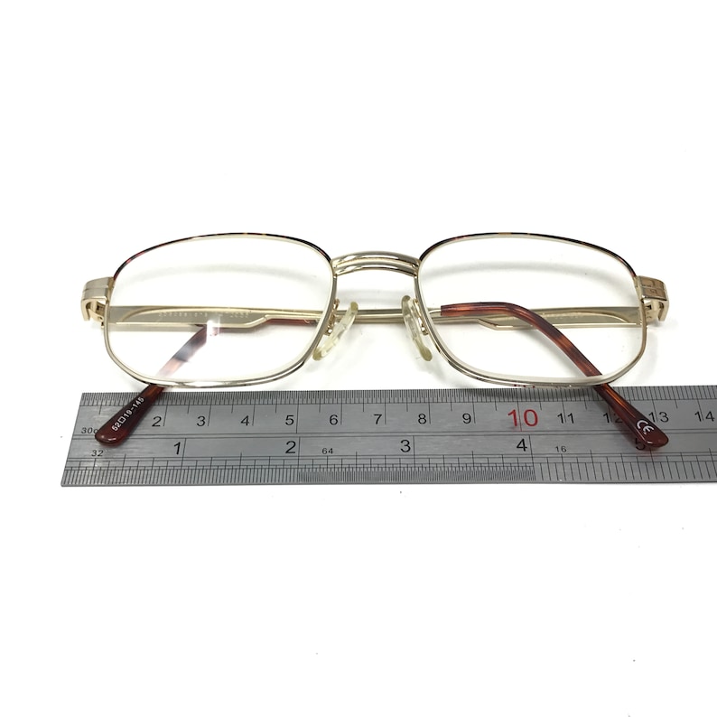 May include: Gold-tone metal eyeglasses with a rectangular frame and clear lenses. The eyeglasses are measured with a ruler, showing a width of approximately 10 centimeters.