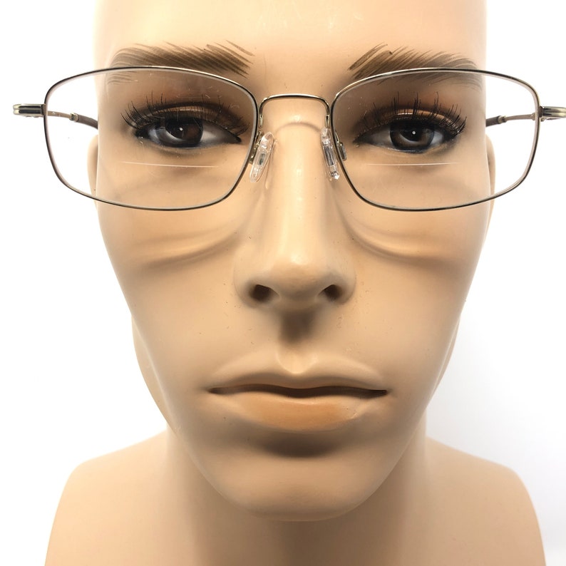 May include: Silver-framed eyeglasses with a rectangular shape are displayed on a mannequin. The glasses have a thin, metallic frame and clear lenses. The nose pads are transparent. The mannequin's face is in focus, with the glasses centered.