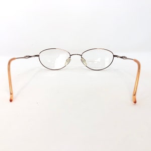 May include: Oval-shaped eyeglasses with a thin, dark brown metal frame. The lenses are clear, and the temples are a light brown color. The glasses are set against a white background.