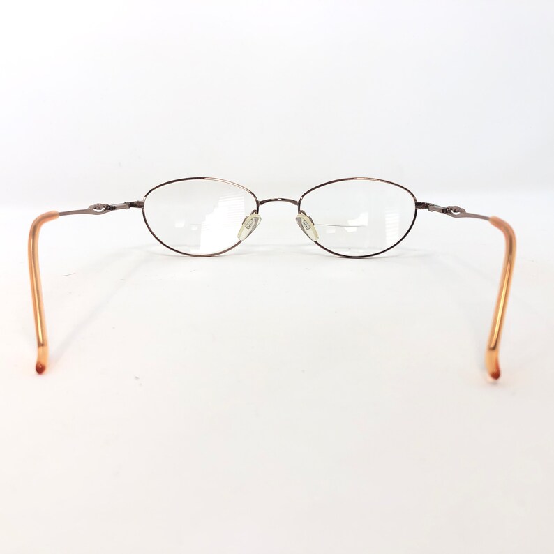 May include: Oval-shaped eyeglasses with a thin, dark brown metal frame. The lenses are clear, and the temples are a light brown color. The glasses are set against a white background.
