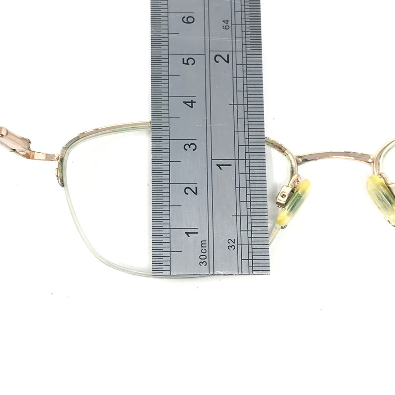 May include: Close-up of a pair of eyeglasses with a silver frame. The glasses are being measured with a ruler, showing a length of approximately 2 inches. The lenses are clear, and the nose pads are yellow.