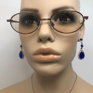 May include: A mannequin head wearing brown oval-framed eyeglasses with tinted lenses and dangling blue teardrop earrings. The earrings have a silver-colored setting and are adorned with clear crystals and a large blue stone.