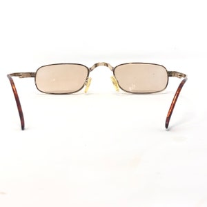 May include: Rectangular sunglasses with a light brown tint. The frames are a silver color with brown and gold-colored arms. The glasses are set against a white background.