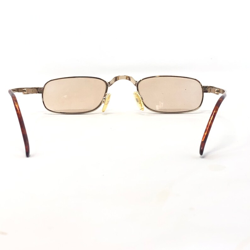 May include: Rectangular sunglasses with a light brown tint. The frames are a silver color with brown and gold-colored arms. The glasses are set against a white background.