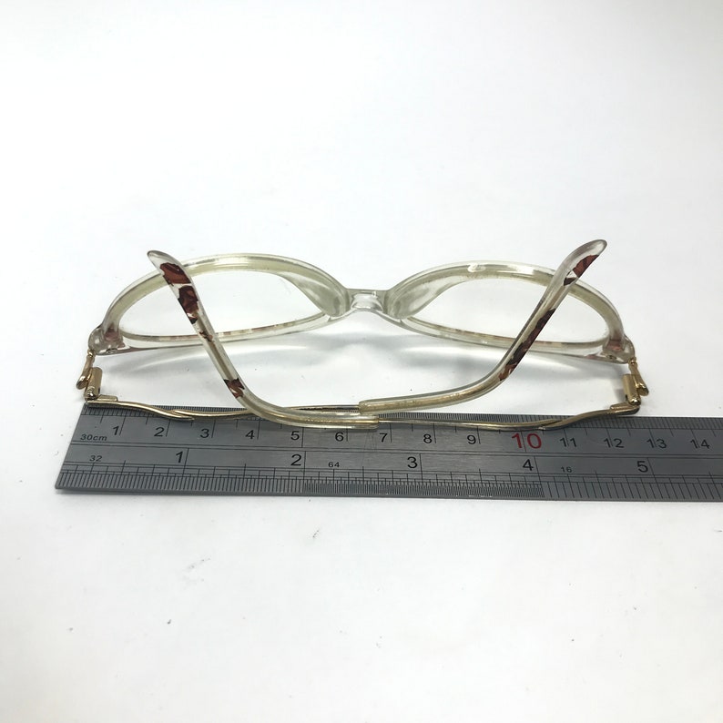 May include: A pair of clear plastic glasses with brown and gold accents. The glasses are measured with a ruler, showing the length of the arms is 10 cm.