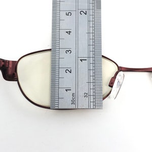 May include: Close-up of a pair of maroon-colored eyeglasses with a ruler placed over the lens. The ruler shows a measurement of approximately 2 inches. The glasses have a classic oval shape.