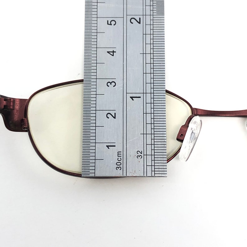 May include: Close-up of a pair of maroon-colored eyeglasses with a ruler placed over the lens. The ruler shows a measurement of approximately 2 inches. The glasses have a classic oval shape.