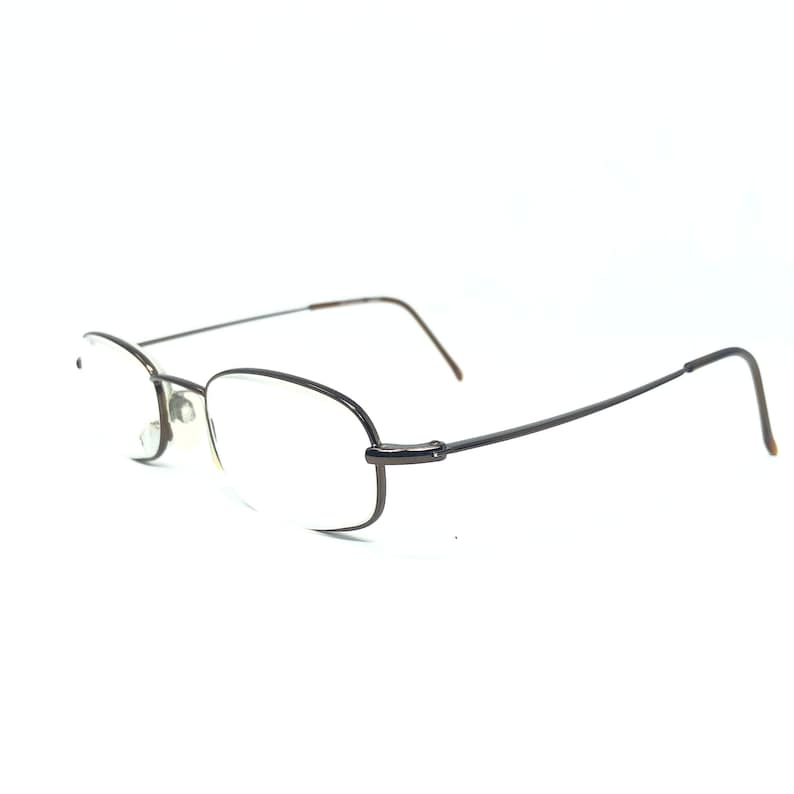 Puede incluir: Un par de gafas con lentes ovaladas y montura de metal marr&oacute;n oscuro. Las gafas tienen una barra superior fina y recta y patillas que se curvan hacia la oreja. Las lentes son transparentes.