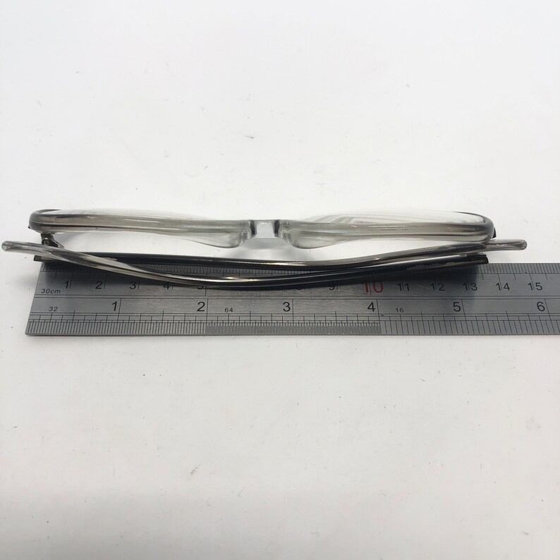K&ouml;nnte beinhalten: Brille mit transparentem Rahmen und schwarzem B&uuml;gel liegt auf einem Lineal. Das Lineal zeigt Messungen in Zentimetern an. Die Brille ist etwa 14 cm breit.