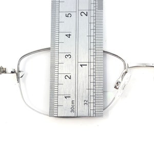May include: Silver-framed eyeglasses with a ruler placed over the lens. The ruler shows measurements in inches and centimeters. The glasses have a rectangular shape and clear nose pads. The frame is thin and metallic.