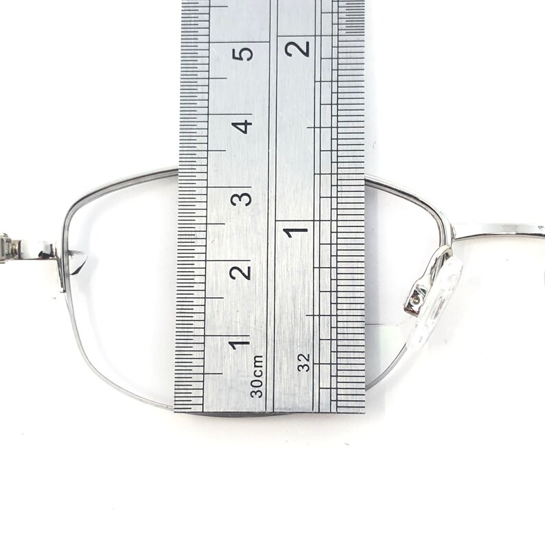 May include: Silver-framed eyeglasses with a ruler placed over the lens. The ruler shows measurements in inches and centimeters. The glasses have a rectangular shape and clear nose pads. The frame is thin and metallic.
