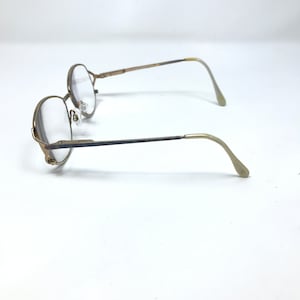 Op de afbeelding: Een paar vintage brillen met ronde, goudkleurige monturen en heldere glazen. De veren zijn donkerder van kleur en gaan over in een lichter goud bij de ooreinden. De bril staat tegen een witte achtergrond.