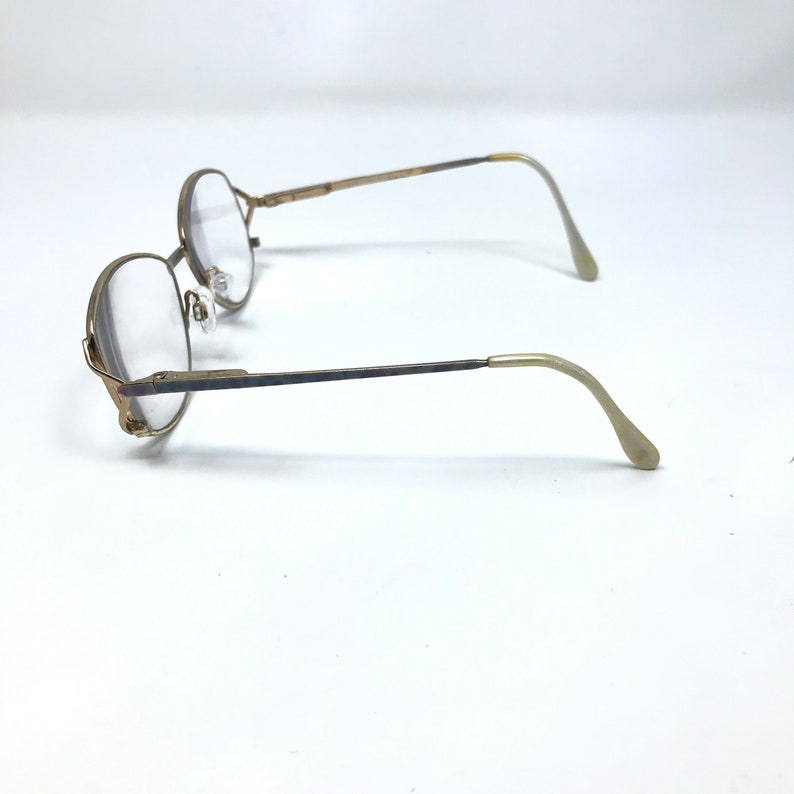 Op de afbeelding: Een paar vintage brillen met ronde, goudkleurige monturen en heldere glazen. De veren zijn donkerder van kleur en gaan over in een lichter goud bij de ooreinden. De bril staat tegen een witte achtergrond.