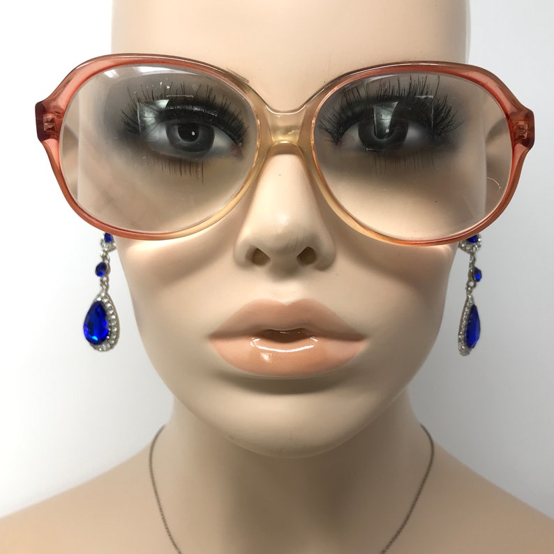 May include: A mannequin wearing large, round, peach-colored glasses with clear lenses. The mannequin is also wearing blue teardrop-shaped earrings with silver accents.